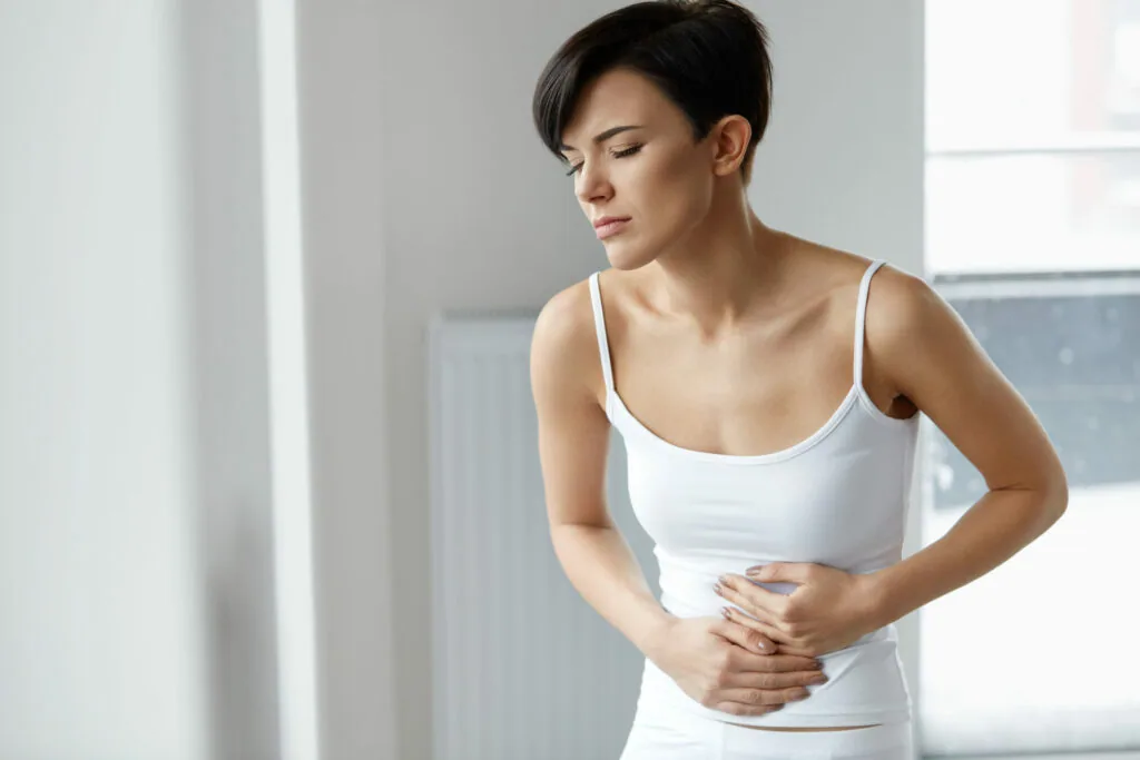 Woman in white tank top holding her stomach in discomfort while looking down
