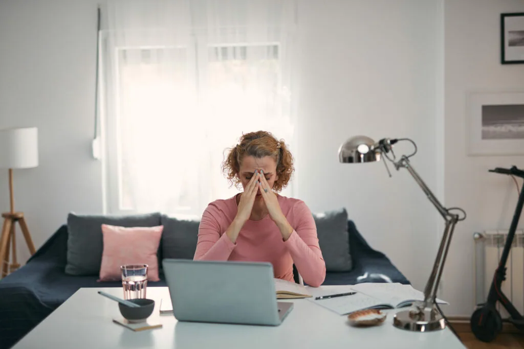 Woman with reddish-brown curly hair in pink top sitting at desk with laptop, hands on face showing fatigue or stress, in modern home office with dark sofa and desk lamp