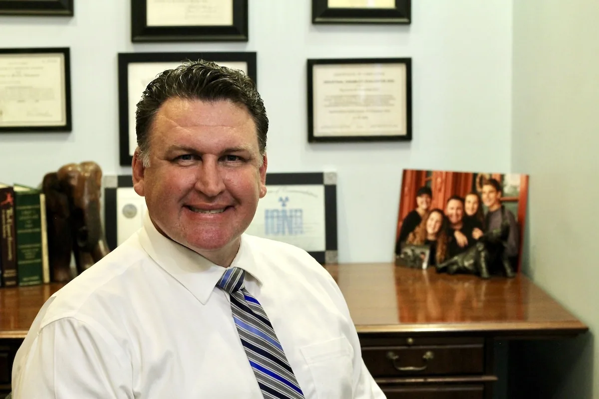 Man in white shirt and blue striped tie smiling at desk in office with framed certificates on wall