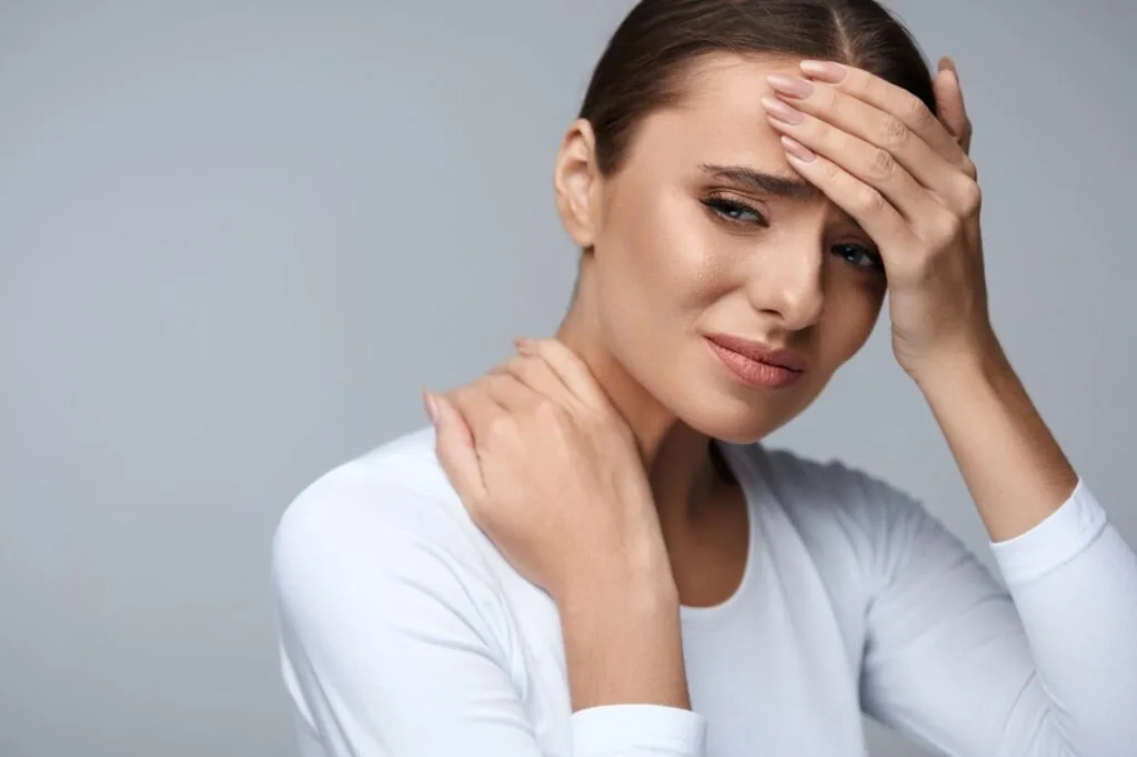 Woman in white long-sleeve shirt holding her forehead and neck with hands, appearing to experience pain or discomfort