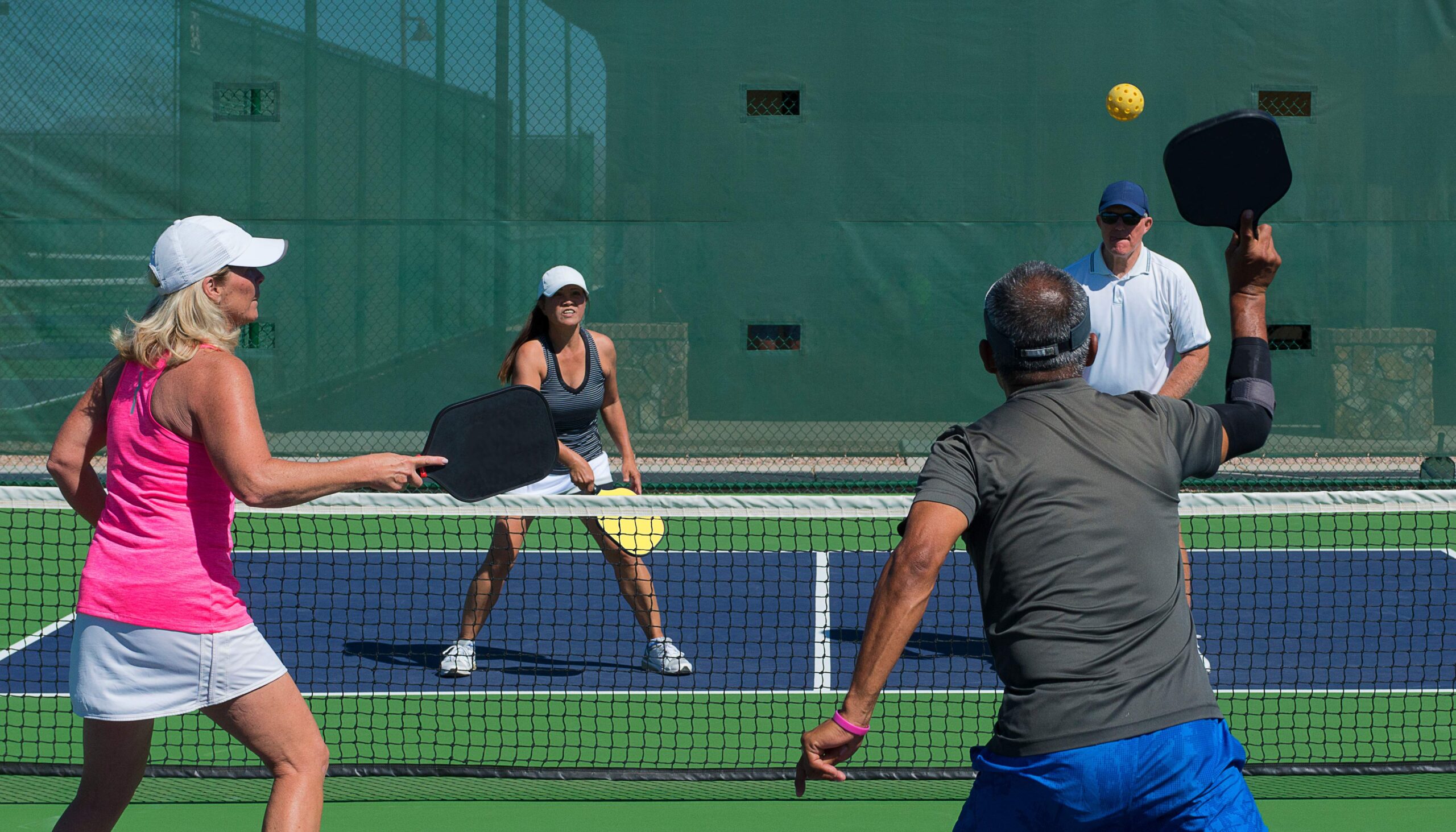 Four people playing doubles pickleball on an outdoor court with green wind screens and blue net